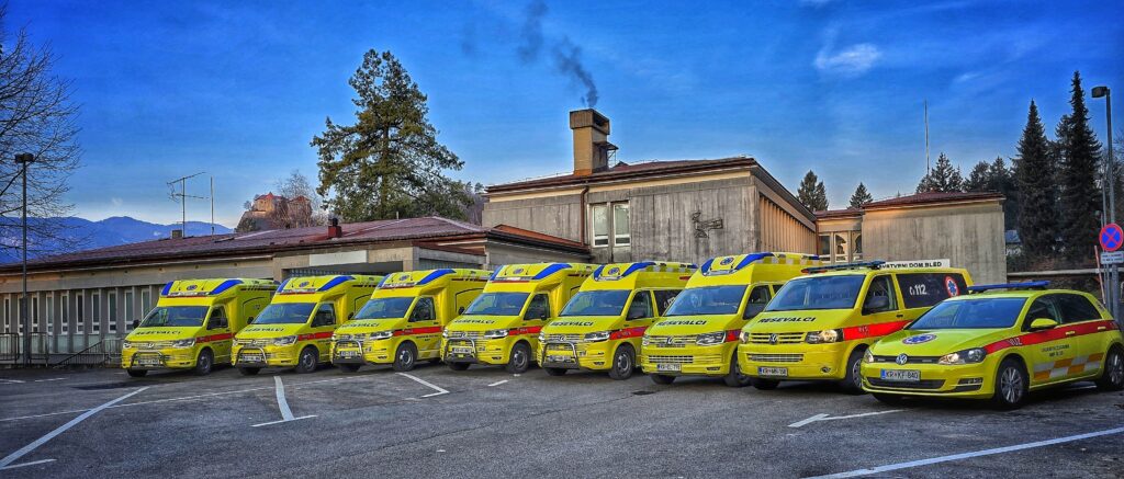 Sedem rumenih reševalnih vozil nujne medicinske pomoči parkiranih pred stavbo, v ozadju drevesa in gore.